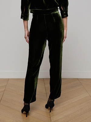 A person stands on a wooden floor, facing away, wearing L'AGENCE Rebel Velvet Trouser in olive with a matching top and black heeled shoes. The outfit features a soft, textured look.