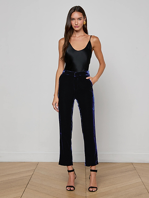 A woman with long brown hair stands against a plain wall, wearing L'AGENCE Rebel Velvet Trouser in midnight blue, a black sleeveless top, and black high-heeled sandals, with one hand resting in her pocket.