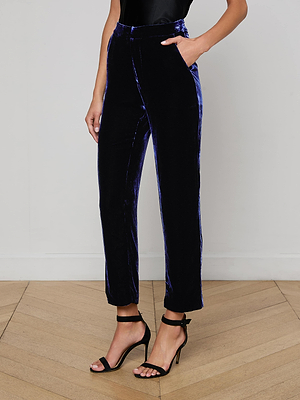 A person wears L'AGENCE's Rebel Velvet Trouser in midnight blue—high-rise, straight-leg style—paired with a black sleeveless top and black strappy heeled sandals, standing on a light wood floor against a white wall.