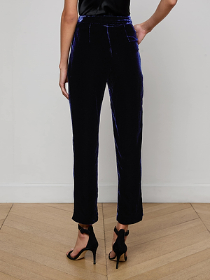 A person stands on a wooden floor wearing L'AGENCE Rebel Velvet Trouser in midnight blue with black high-heeled sandals. The photo, taken from behind, highlights the trouser’s fit and lower back.