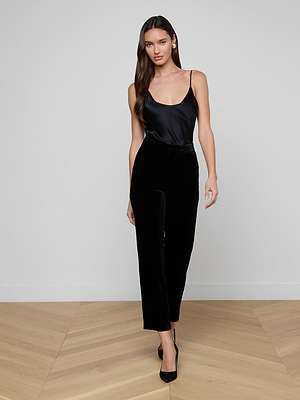 A woman with long brown hair wears a black satin camisole, the Rebel Velvet Trouser, and black heels, standing on a light wooden floor against a plain white wall.