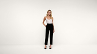A woman stands against a plain white background, wearing a white sleeveless top, black high-waisted pants, and black heels. She has one hand in her pocket and is looking at the camera.