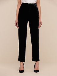 Wearing the Rebel Velvet Trouser, a black sleeveless top, and pointed-toe heels, a person stands on a light wood chevron floor against a plain wall with hands casually in their pockets.