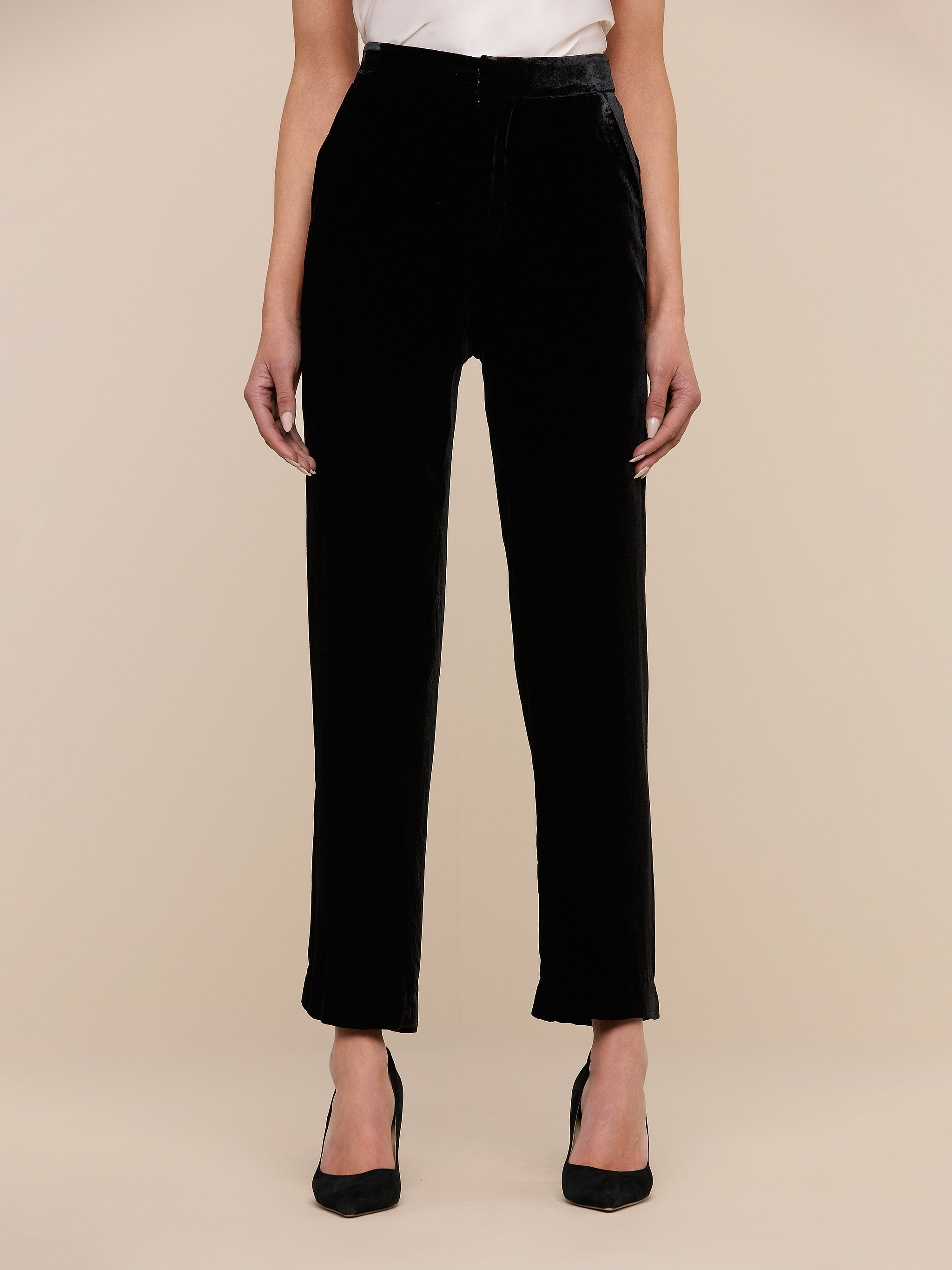 Wearing the Rebel Velvet Trouser, a black sleeveless top, and pointed-toe heels, a person stands on a light wood chevron floor against a plain wall with hands casually in their pockets.