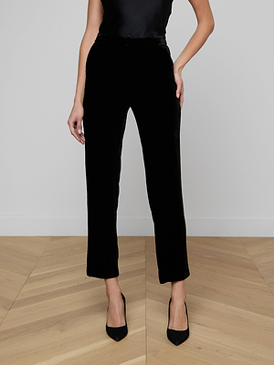 Wearing the Rebel Velvet Trouser, a black sleeveless top, and pointed-toe heels, a person stands on a light wood chevron floor against a plain wall with hands casually in their pockets.