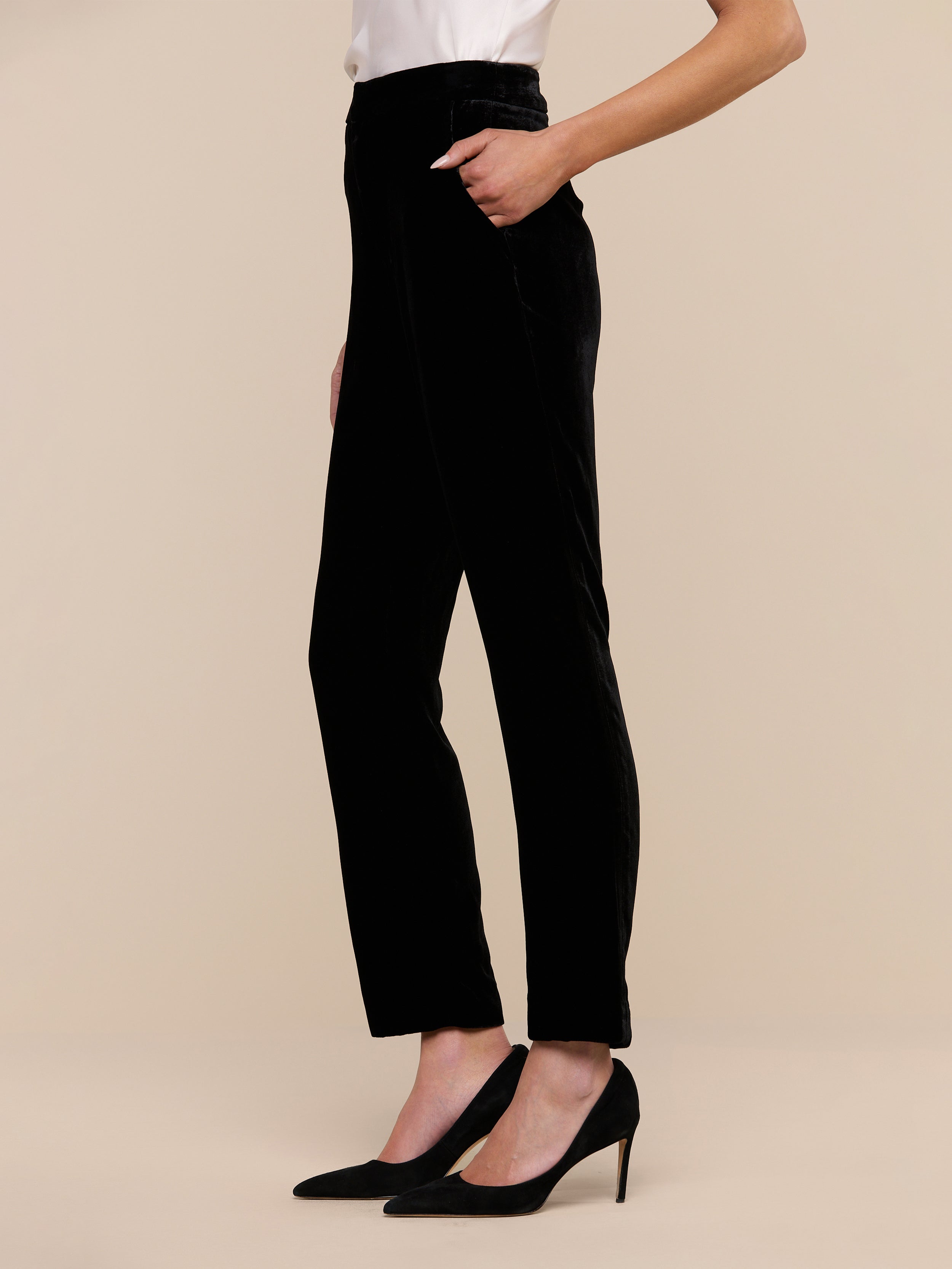 A person wearing the L’AGENCE Rebel Velvet Trouser in black, paired with a black sleeveless top and black pointed-toe heels, stands on a light wood floor with one hand in their pocket. The photo is cropped at the shoulders.