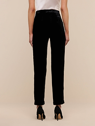 A person stands on a wooden floor facing away from the camera, wearing Rebel Velvet Trouser and black high-heeled shoes against a plain white wall.
