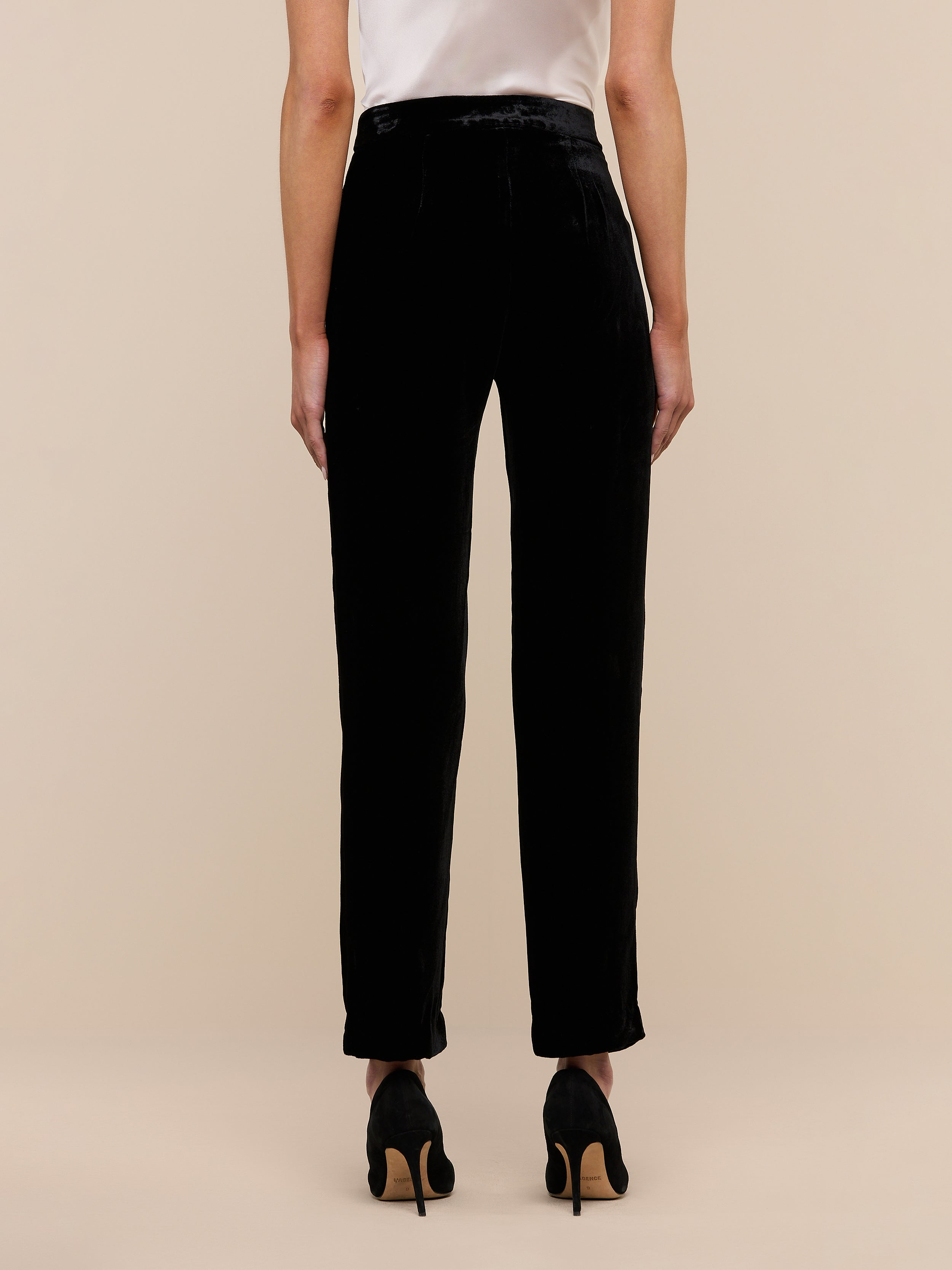 A person stands on a wooden floor facing away from the camera, wearing Rebel Velvet Trouser and black high-heeled shoes against a plain white wall.