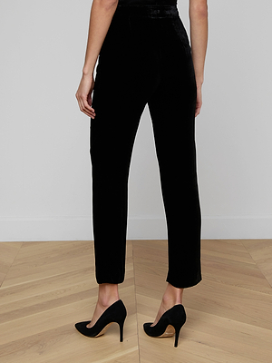 A person stands on a wooden floor facing away from the camera, wearing Rebel Velvet Trouser and black high-heeled shoes against a plain white wall.