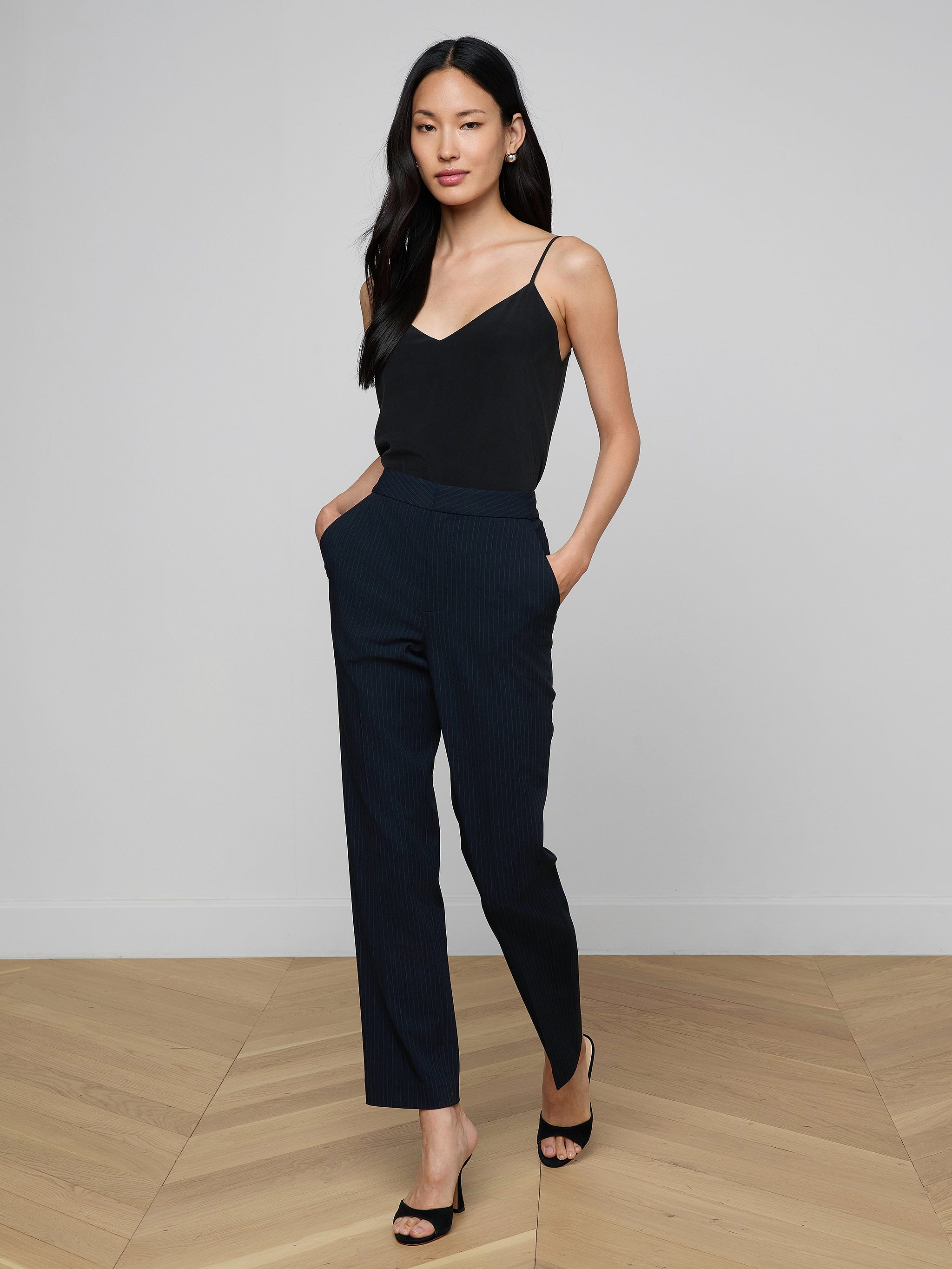 A woman stands with her hands in her pockets, wearing a black sleeveless top, the high-rise Rebel Pinstriped Trouser in navy, and black heels against a plain white background.