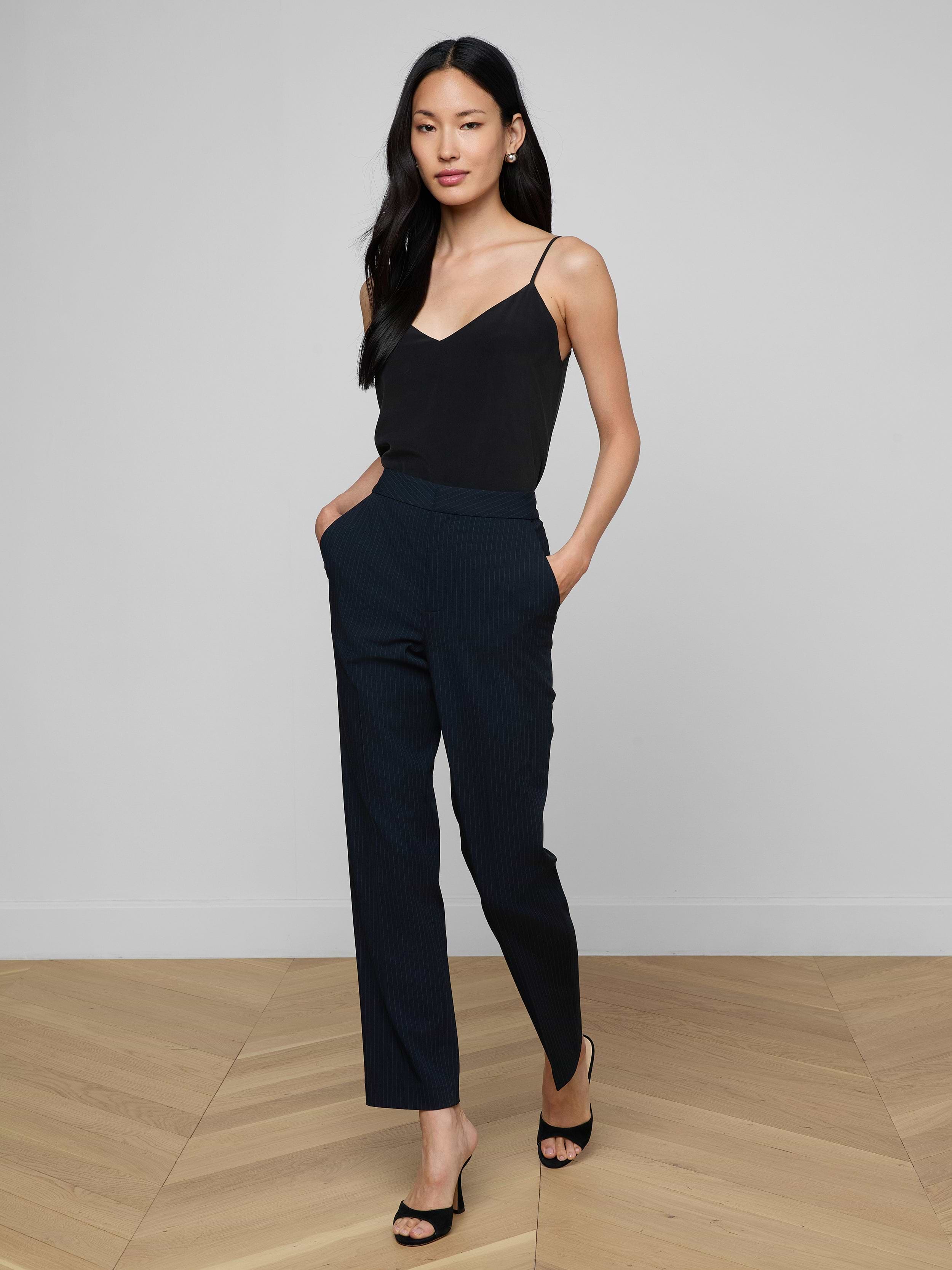 A woman stands with her hands in her pockets, wearing a black sleeveless top, the high-rise Rebel Pinstriped Trouser in navy, and black heels against a plain white background.