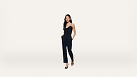 A woman with long black hair wears a black sleeveless top, navy blue pants, and black heels, standing with hands in pockets against a plain white background.