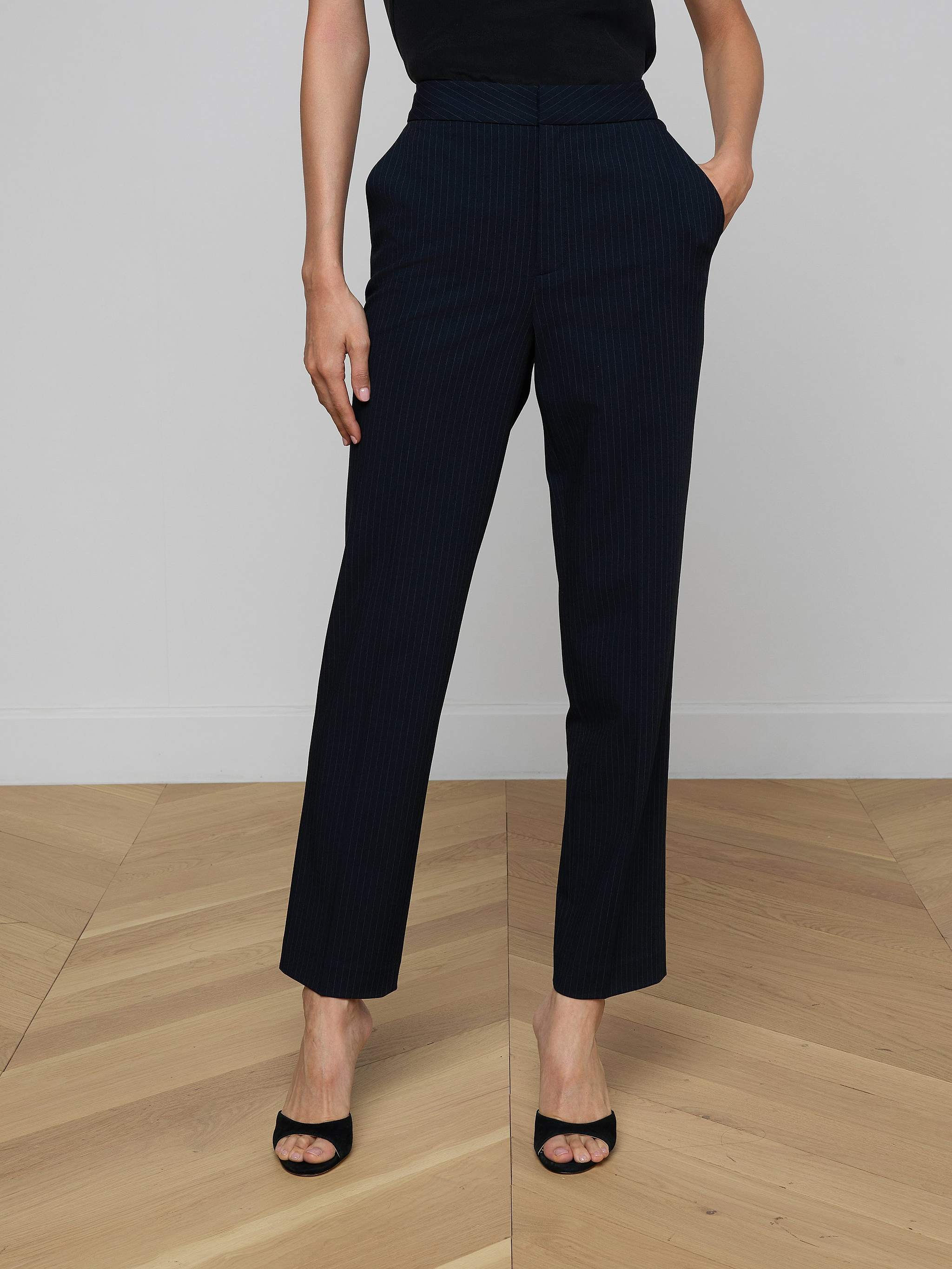 Wearing the Rebel Pinstriped Trouser in black high rise, paired with black open-toe heels and hands casually tucked into pockets, set against a plain light background. Upper body is partially out of frame.