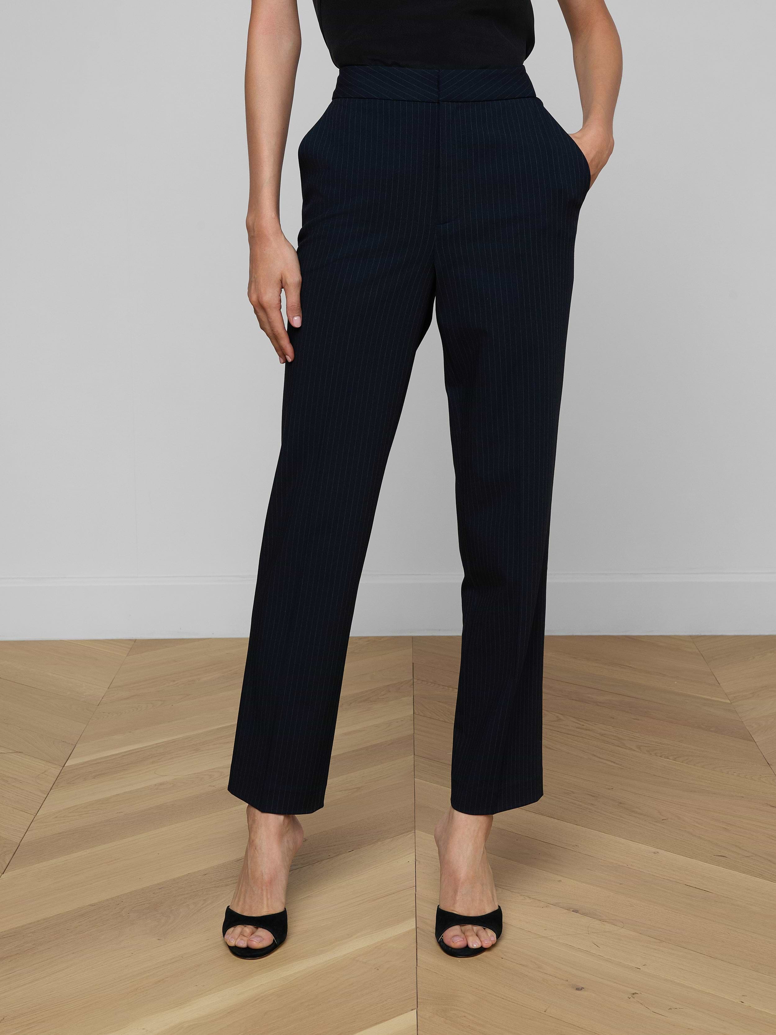 Wearing the Rebel Pinstriped Trouser in black high rise, paired with black open-toe heels and hands casually tucked into pockets, set against a plain light background. Upper body is partially out of frame.