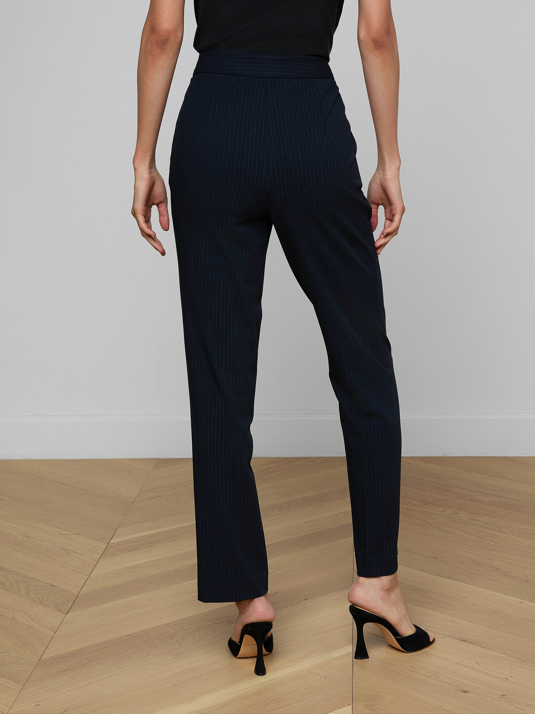 A person wearing the Rebel Pinstriped Trouser, featuring a high-rise, straight-leg silhouette with navy pinstripes, and black heeled shoes stands with their back to the camera against a plain light background.
