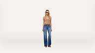 A woman wearing sunglasses, a tan tank top, and blue wide-leg jeans stands facing forward against a plain white background.