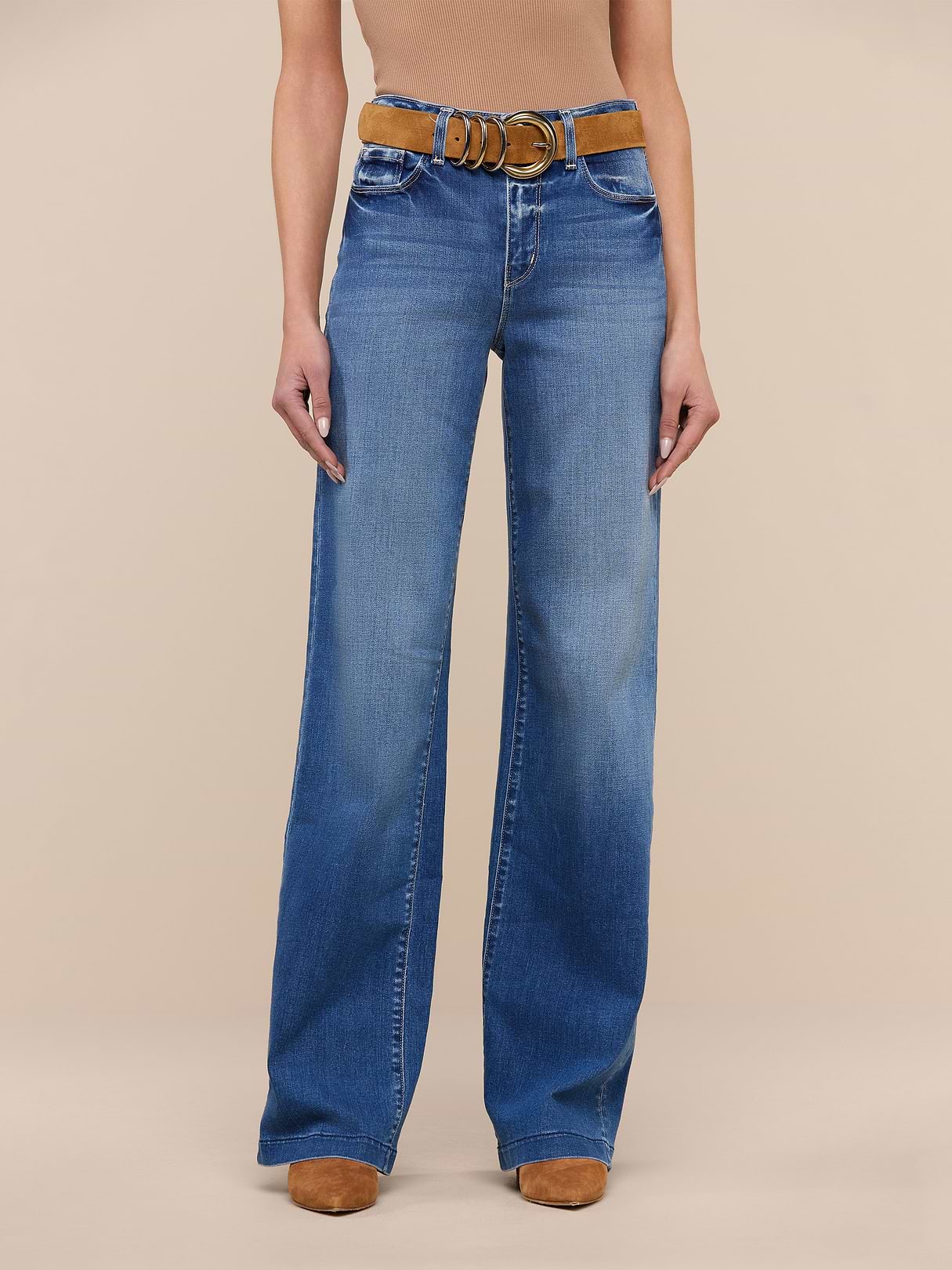 A person stands against a plain white background wearing the Clayton Wide-Leg Jean in high rise denim blue, crafted from REPREVE woven denim, styled with a brown belt and tan shoes. The upper body is cropped out of the image.