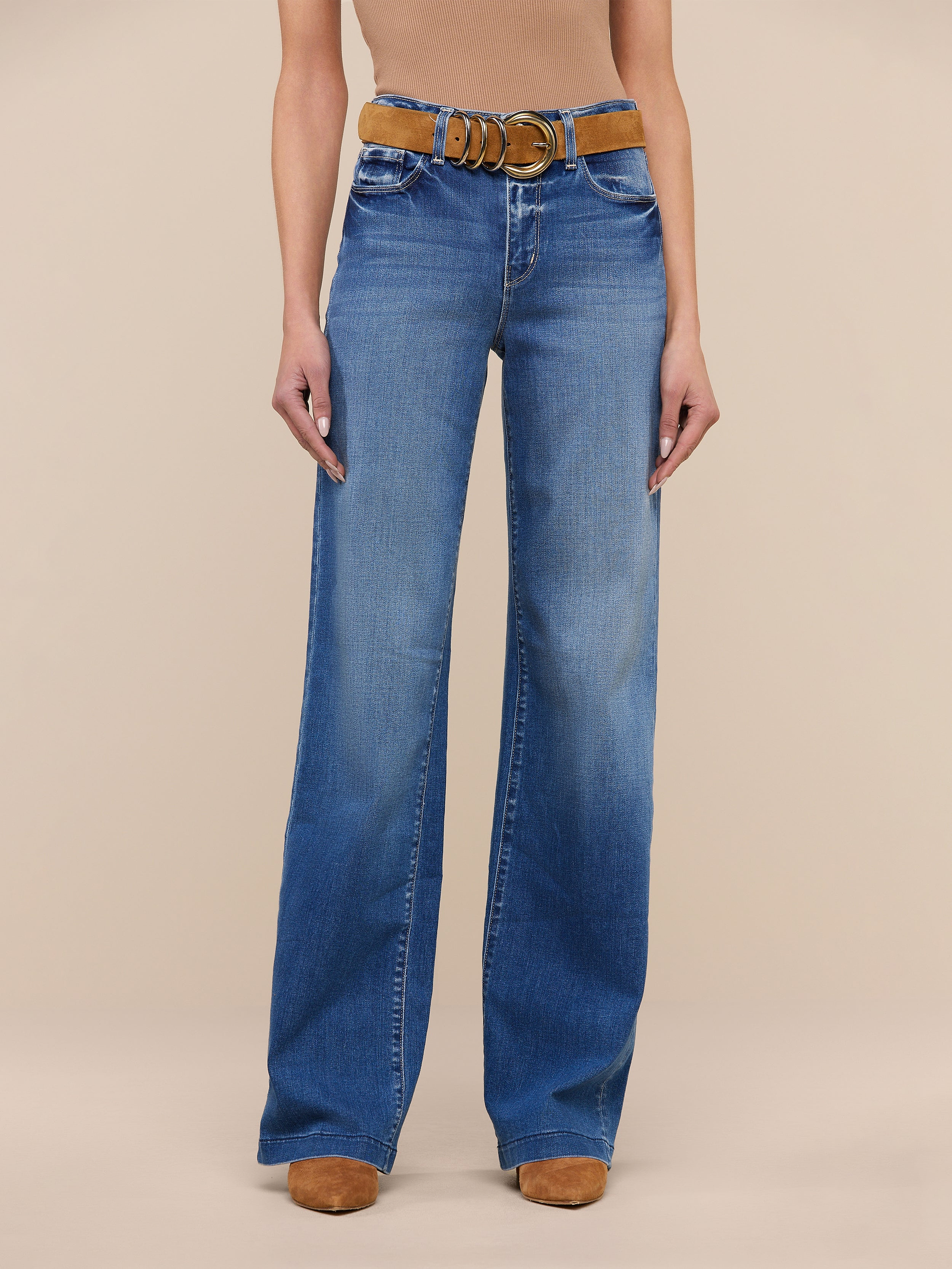 A person stands against a plain white background wearing the Clayton Wide-Leg Jean in high rise denim blue, crafted from REPREVE woven denim, styled with a brown belt and tan shoes. The upper body is cropped out of the image.
