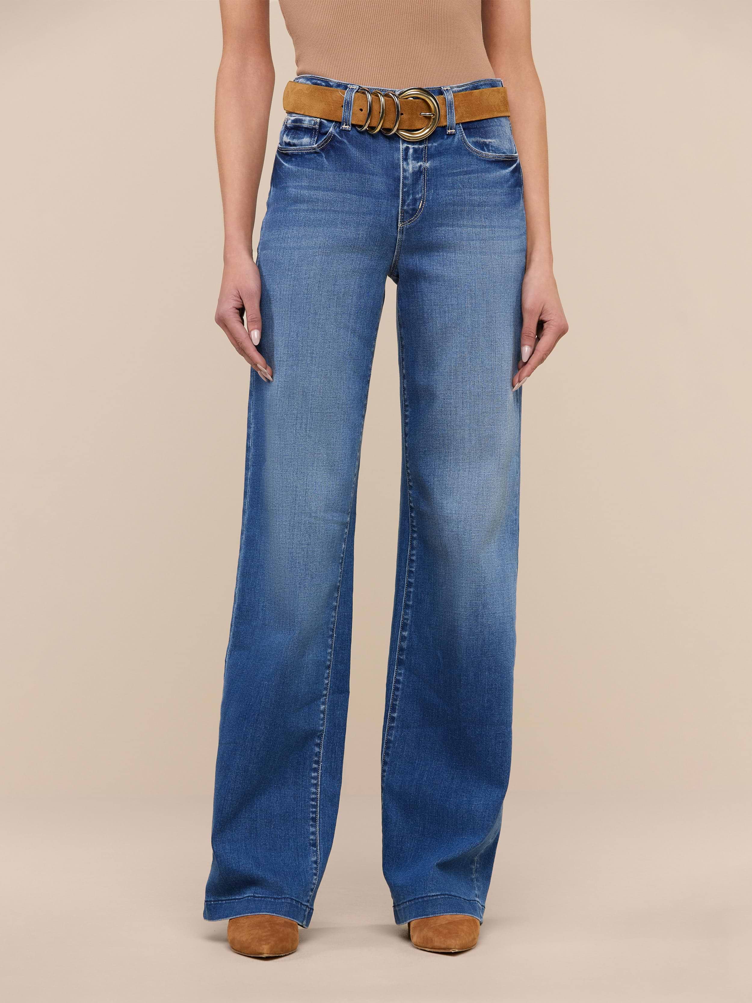 A person stands against a plain white background wearing the Clayton Wide-Leg Jean in high rise denim blue, crafted from REPREVE woven denim, styled with a brown belt and tan shoes. The upper body is cropped out of the image.