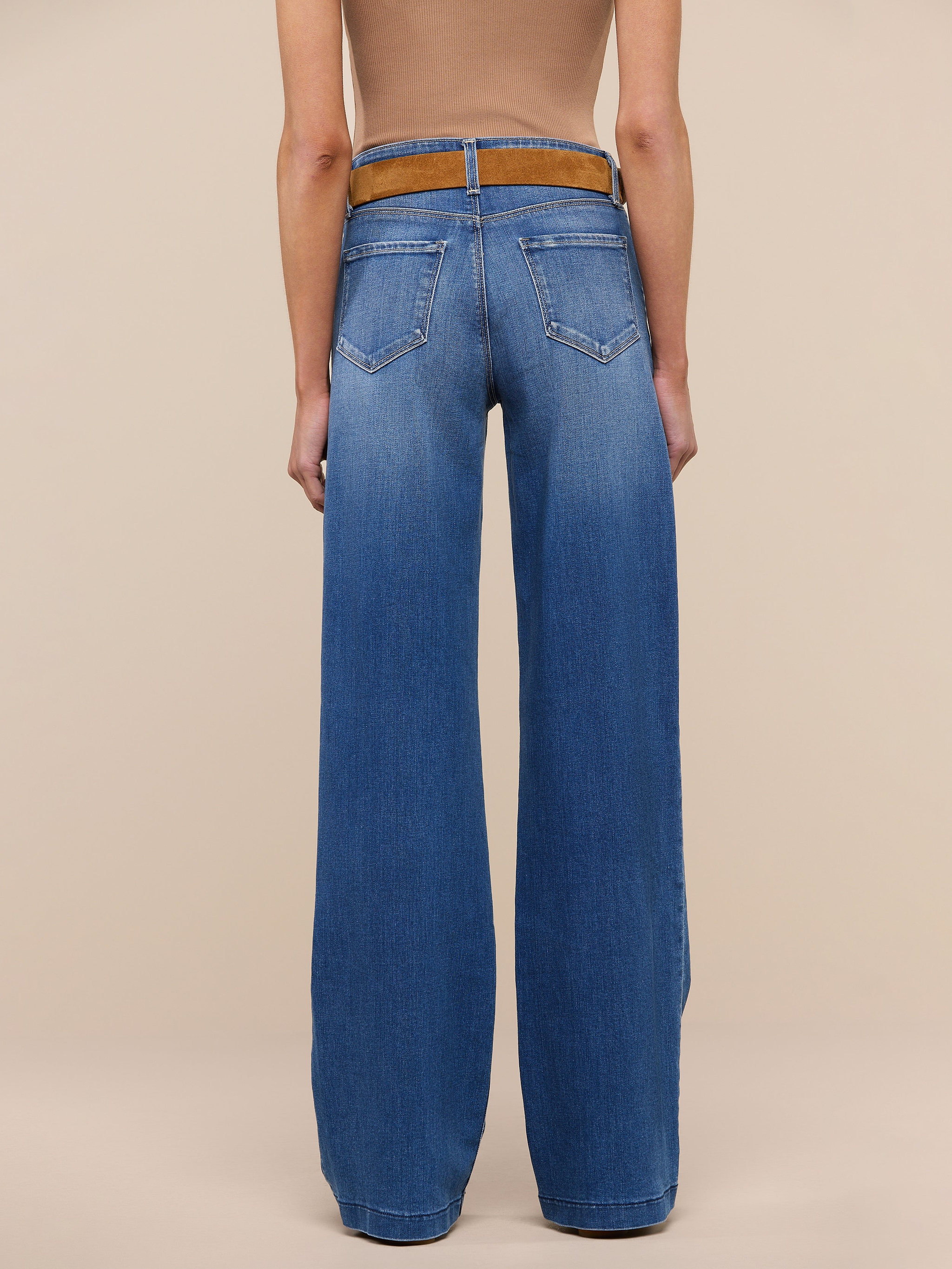 A person stands facing away against a plain white background, wearing a tan sleeveless top with a brown belt and tall wide-leg blue Clayton Wide-Leg Jeans made from REPREVE woven denim.