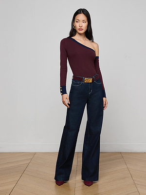 A woman stands indoors facing forward with arms relaxed, wearing the L'AGENCE Clayton Wide-Leg Jean in stretch denim, a burgundy one-shoulder long-sleeve top with navy cuffs, a belt, and burgundy pointed-toe shoes.