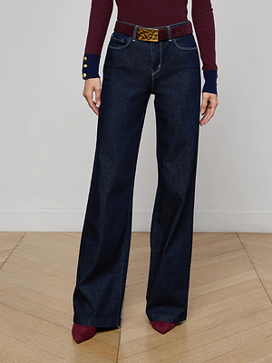 A person wearing L'AGENCE Clayton Wide-Leg Jeans in dark blue stretch denim, a burgundy long-sleeve top with navy cuffs and gold buttons, a patterned belt, and burgundy pointed shoes stands on a wooden floor.