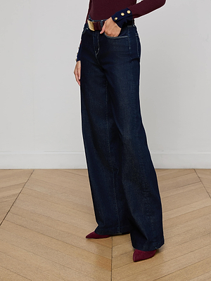 Someone stands on a light wood chevron floor wearing L'AGENCE Clayton Wide-Leg Jeans in dark blue stretch denim, paired with a burgundy top featuring gold buttons and matching burgundy pointed-toe shoes.