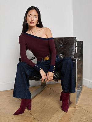 A woman sits on a black chair wearing a burgundy one-shoulder top, L’AGENCE Clayton Wide-Leg Jean in stretch denim, burgundy boots, gold hoop earrings, with her hair down and arms resting on her knees.