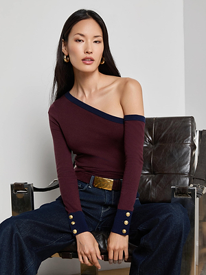 A woman with straight dark hair sits on a leather chair, wearing the L'AGENCE Clayton Wide-Leg Jean, a burgundy one-shoulder top with navy trim and gold buttons, gold earrings, and a wide brown belt with a gold buckle.