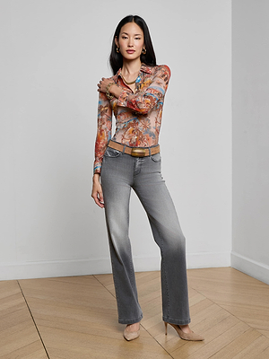 A woman stands in a room with wooden floors, wearing the Clayton Wide-Leg Jean in vintage gray wash, styled with a colorful long-sleeve top, brown belt, nude heels, and gold jewelry. She poses with one hand touching her shoulder.