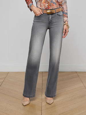 Someone stands on a wooden floor against a plain light wall wearing Clayton Wide-Leg Jeans in vintage gray wash, a brown belt, a patterned long-sleeve top, and beige pointed-toe heels.