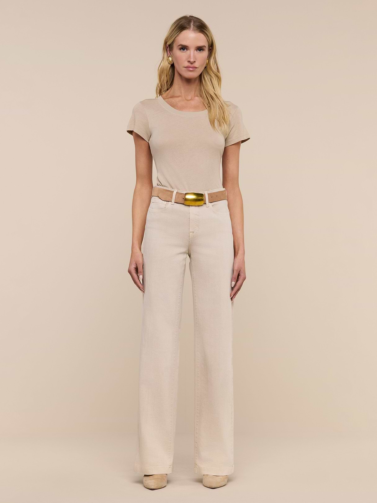 A woman stands facing forward against a beige background, wearing a short-sleeved beige top, the Clayton Wide-Leg Jean with a matching belt and gold buckle, and beige shoes.