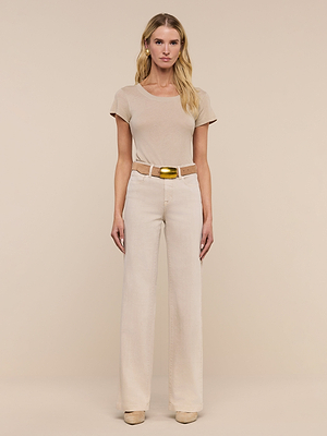 A woman stands facing forward against a beige background, wearing a short-sleeved beige top, the Clayton Wide-Leg Jean with a matching belt and gold buckle, and beige shoes.