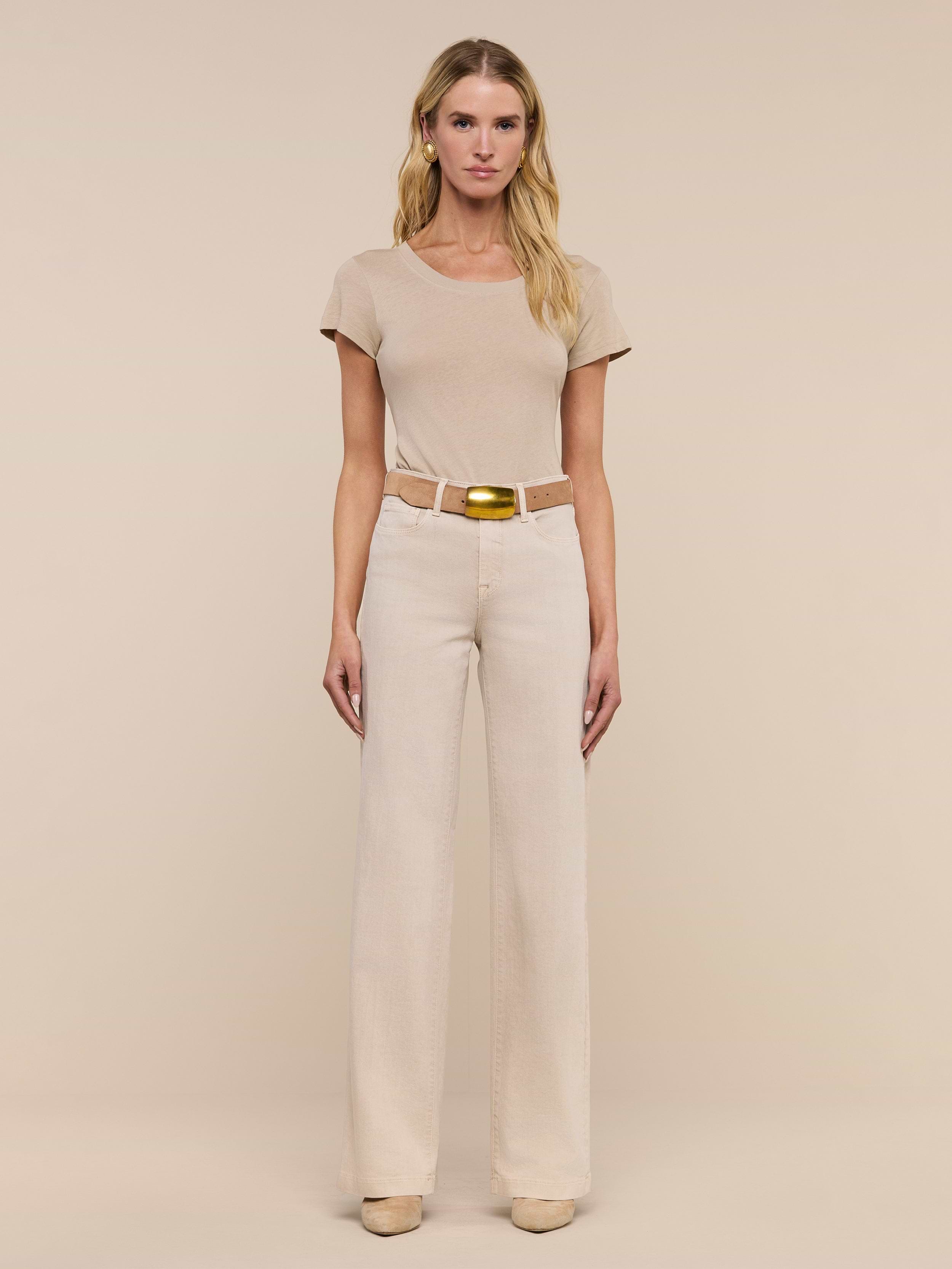 A woman stands facing forward, wearing a beige short-sleeve top, beige shoes, and the Clayton Wide-Leg Jean in a matching high-rise, tall wide-leg style with a belt, against a plain white background.