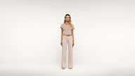 A woman stands facing forward in a cream-colored, short-sleeved top and matching wide-legged pants, accessorized with a gold belt, against a plain white background.