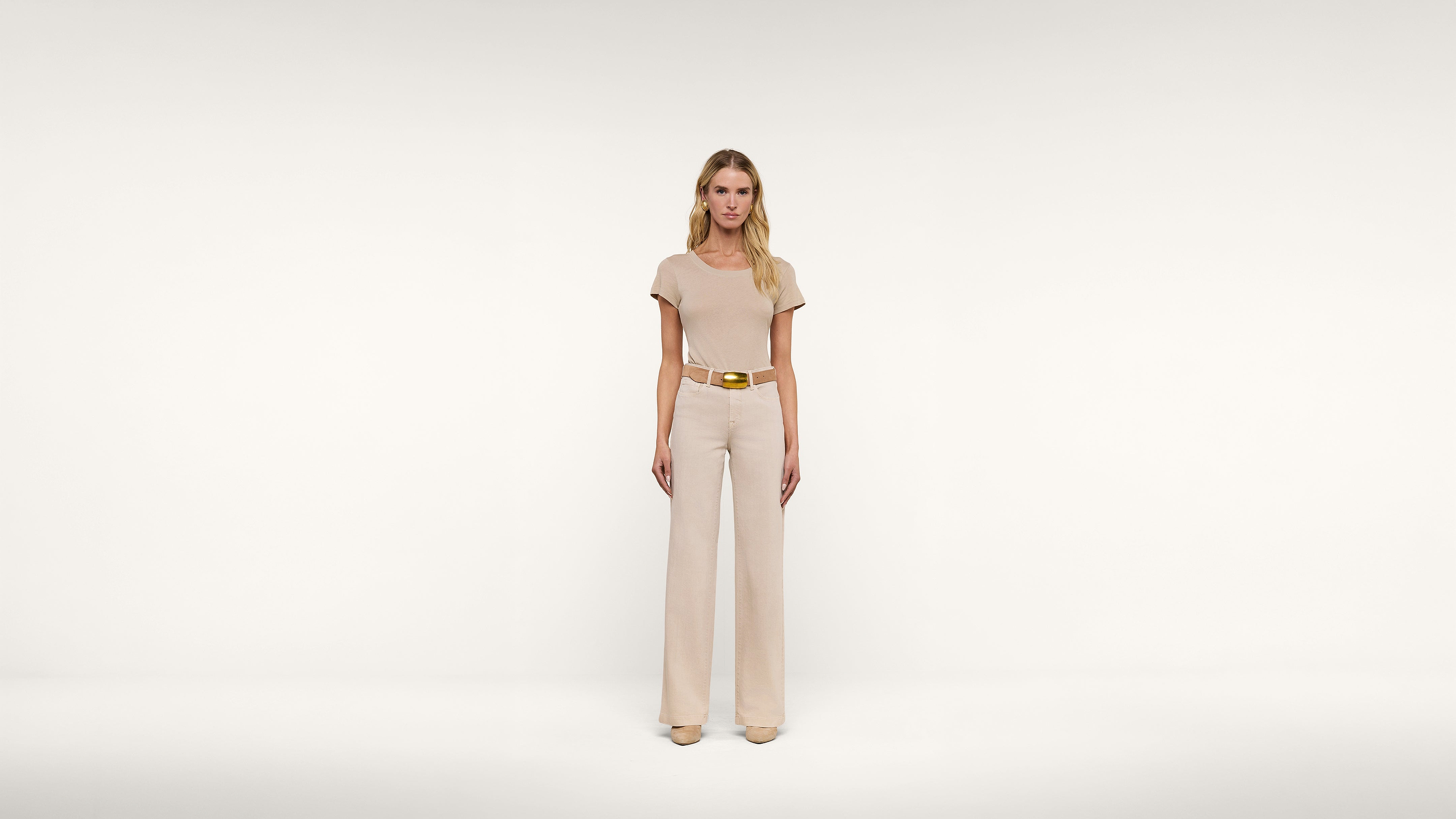 A woman stands facing forward in a cream-colored, short-sleeved top and matching wide-legged pants, accessorized with a gold belt, against a plain white background.