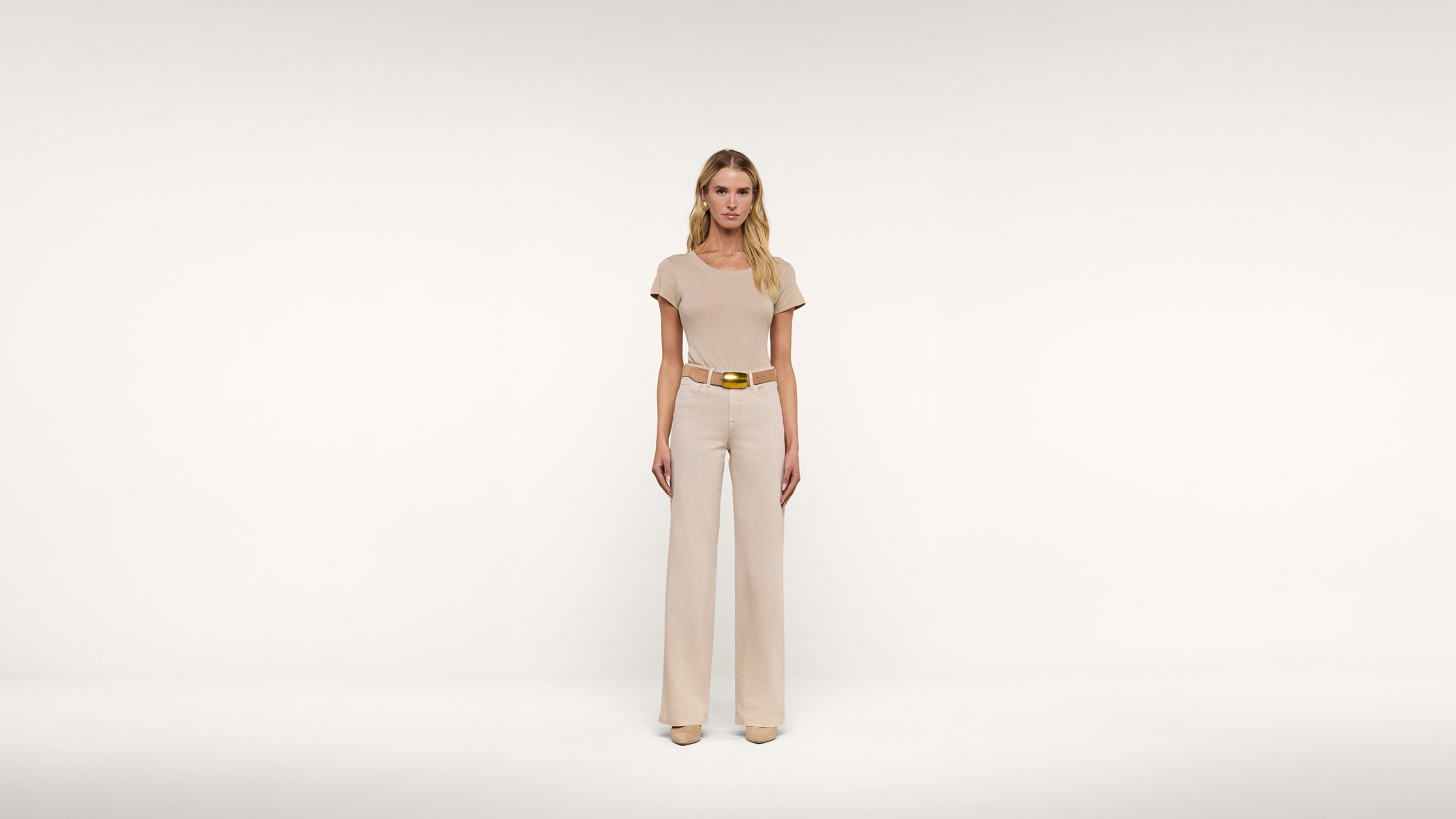 A woman stands facing forward in a cream-colored, short-sleeved top and matching wide-legged pants, accessorized with a gold belt, against a plain white background.