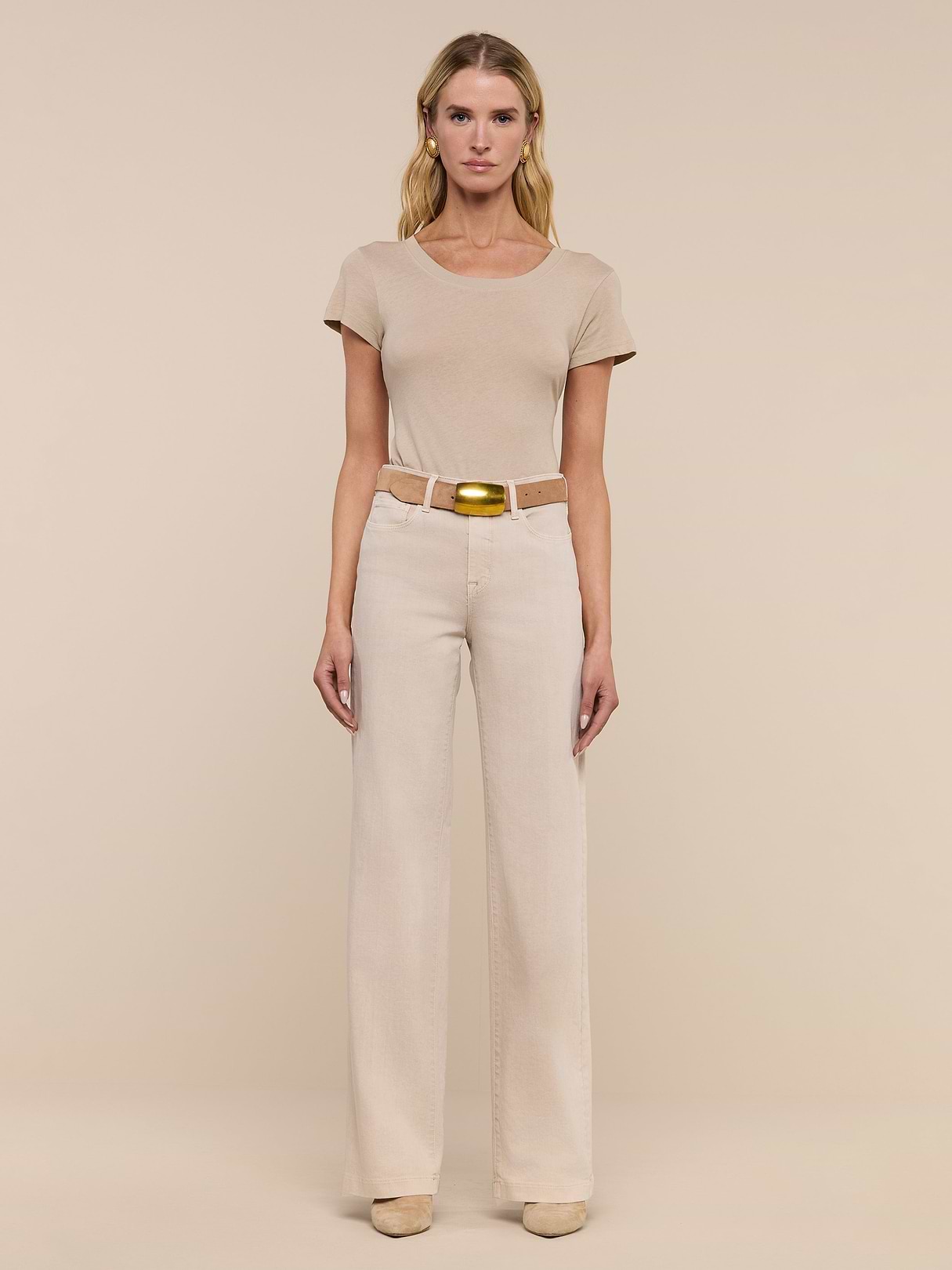A person stands facing forward against a beige background, wearing a short-sleeve beige t-shirt, Clayton Wide-Leg Jean in cream with a beige belt and gold buckle, and beige shoes.