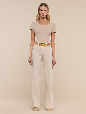 A person stands facing forward against a beige background, wearing a short-sleeve beige t-shirt, Clayton Wide-Leg Jean in cream with a beige belt and gold buckle, and beige shoes.