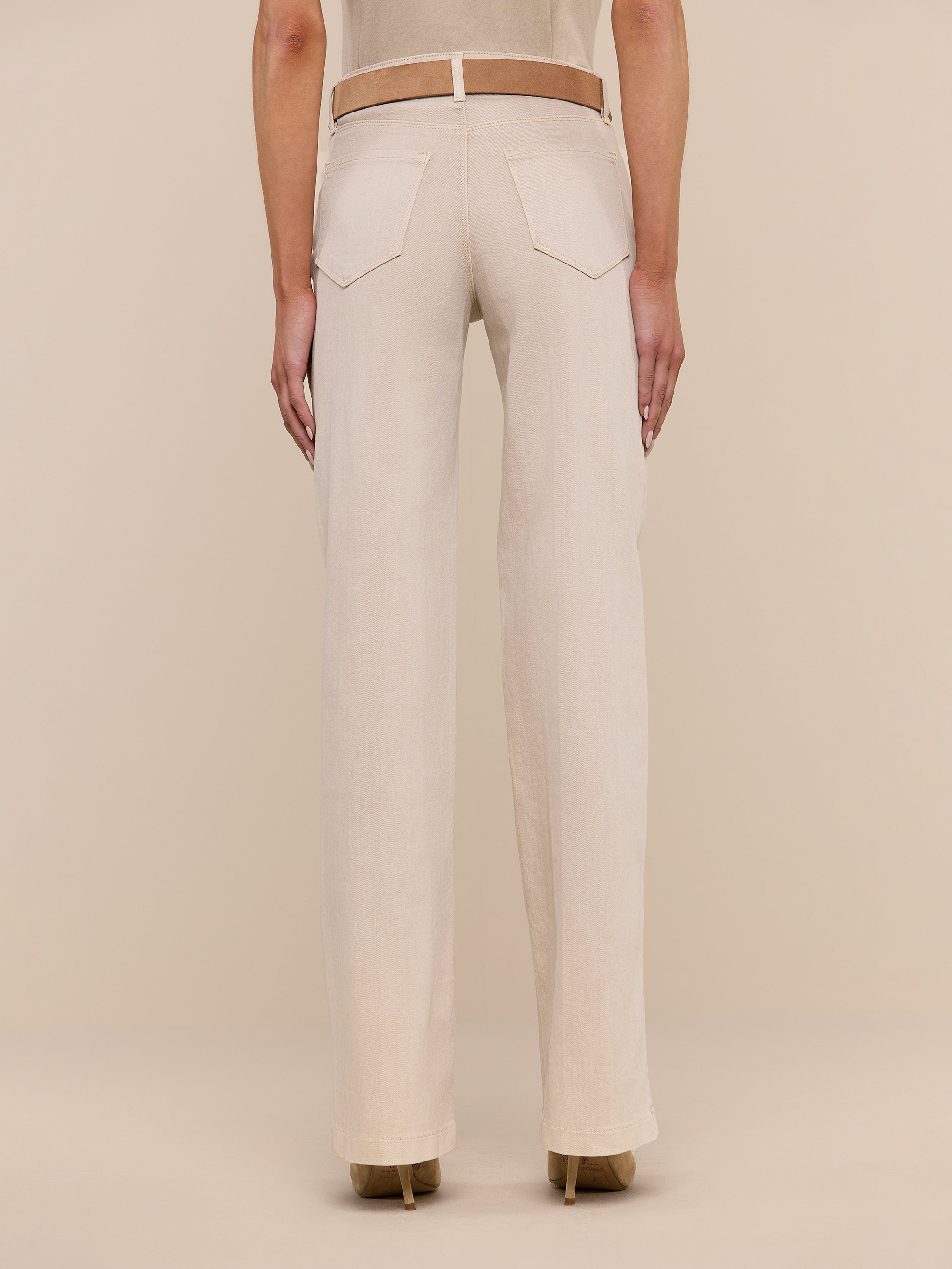 A person is shown from the back, displaying only the lower half of their body, dressed in beige Clayton Wide-Leg Jeans made from stretch denim with back pockets and a fitted waistband. The jeans are paired with a beige top and tan heeled shoes, set against a plain white background.