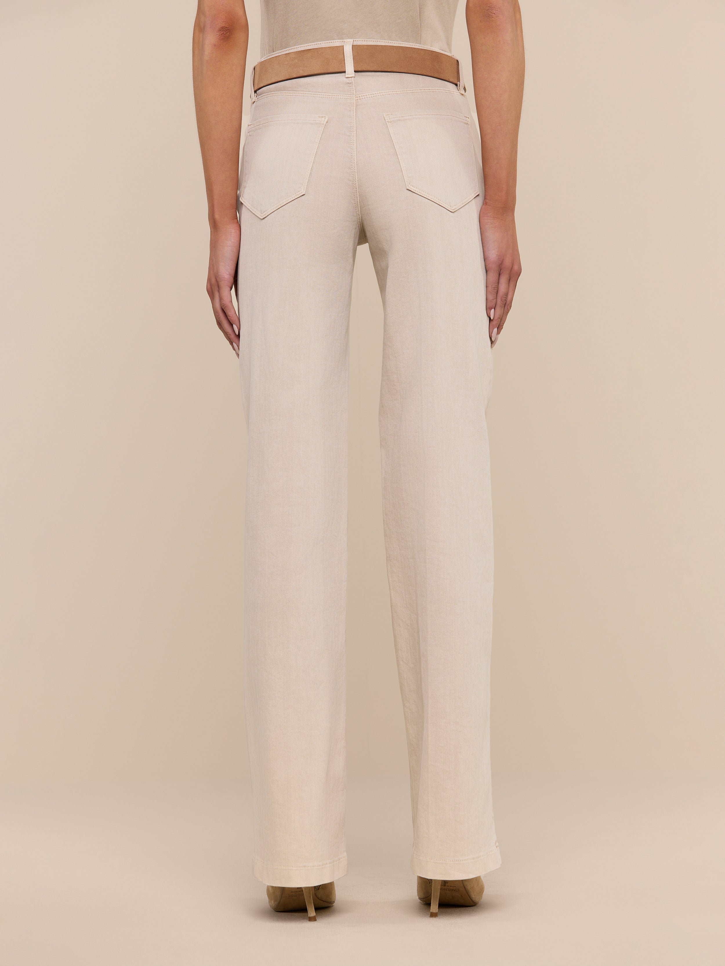 A person is shown from the back, displaying only the lower half of their body, dressed in beige Clayton Wide-Leg Jeans made from stretch denim with back pockets and a fitted waistband. The jeans are paired with a beige top and tan heeled shoes, set against a plain white background.