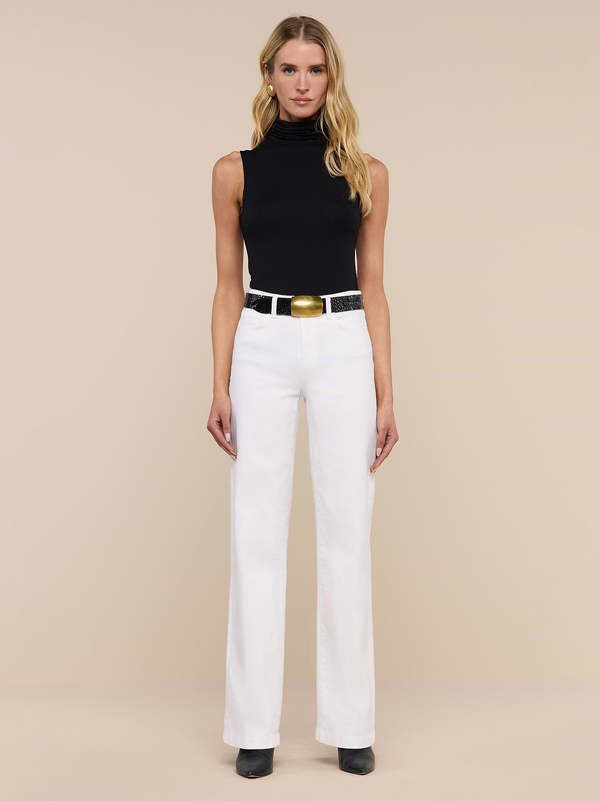 A woman stands facing forward in a black sleeveless turtleneck, Clayton Wide-Leg Jean in white stretch denim with five-pocket design, a black belt with a large gold buckle, and black shoes, against a plain beige background.
