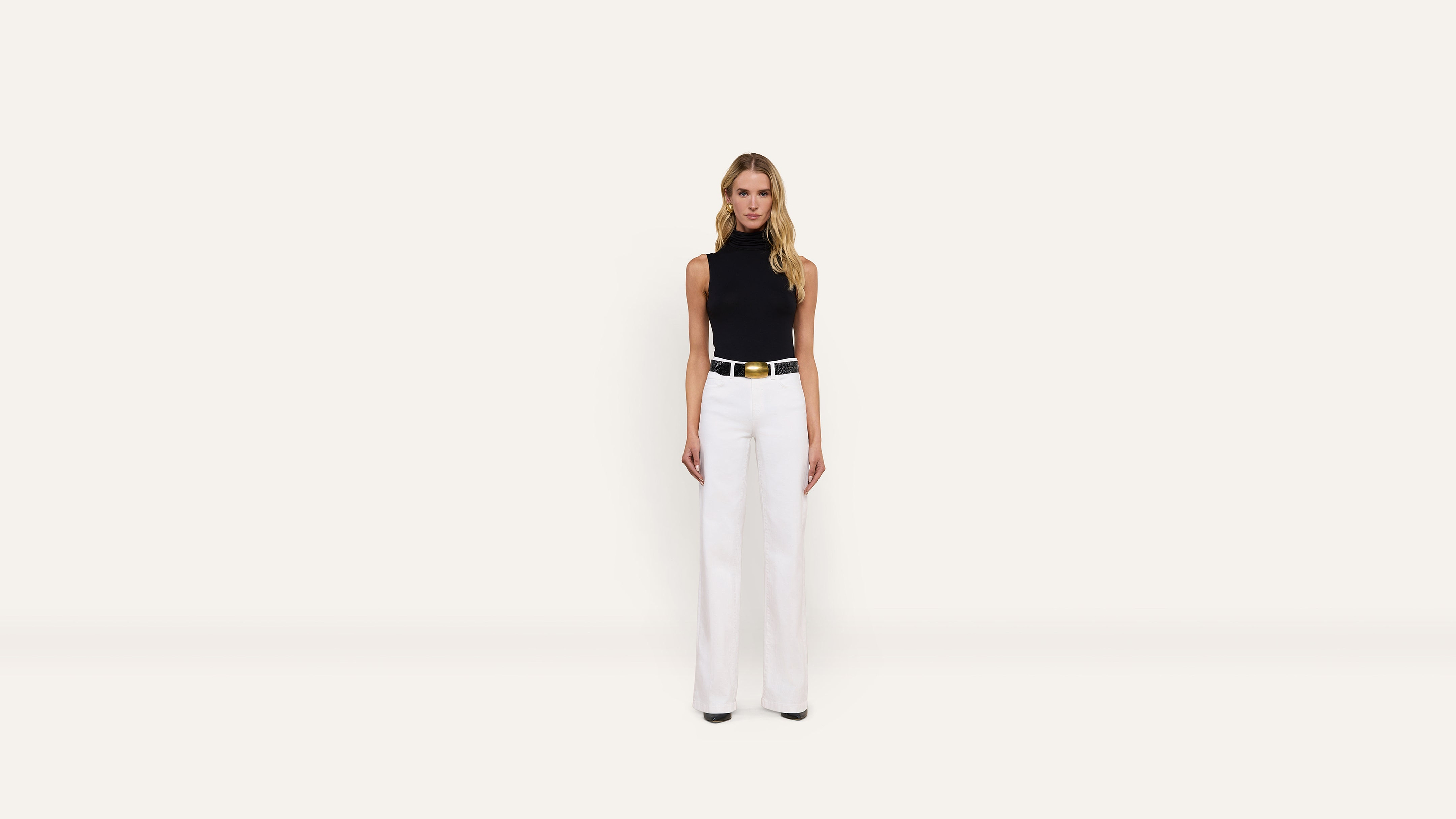 A woman wearing a stylish black sleeveless turtleneck, white trousers, and a black belt with a gold buckle.