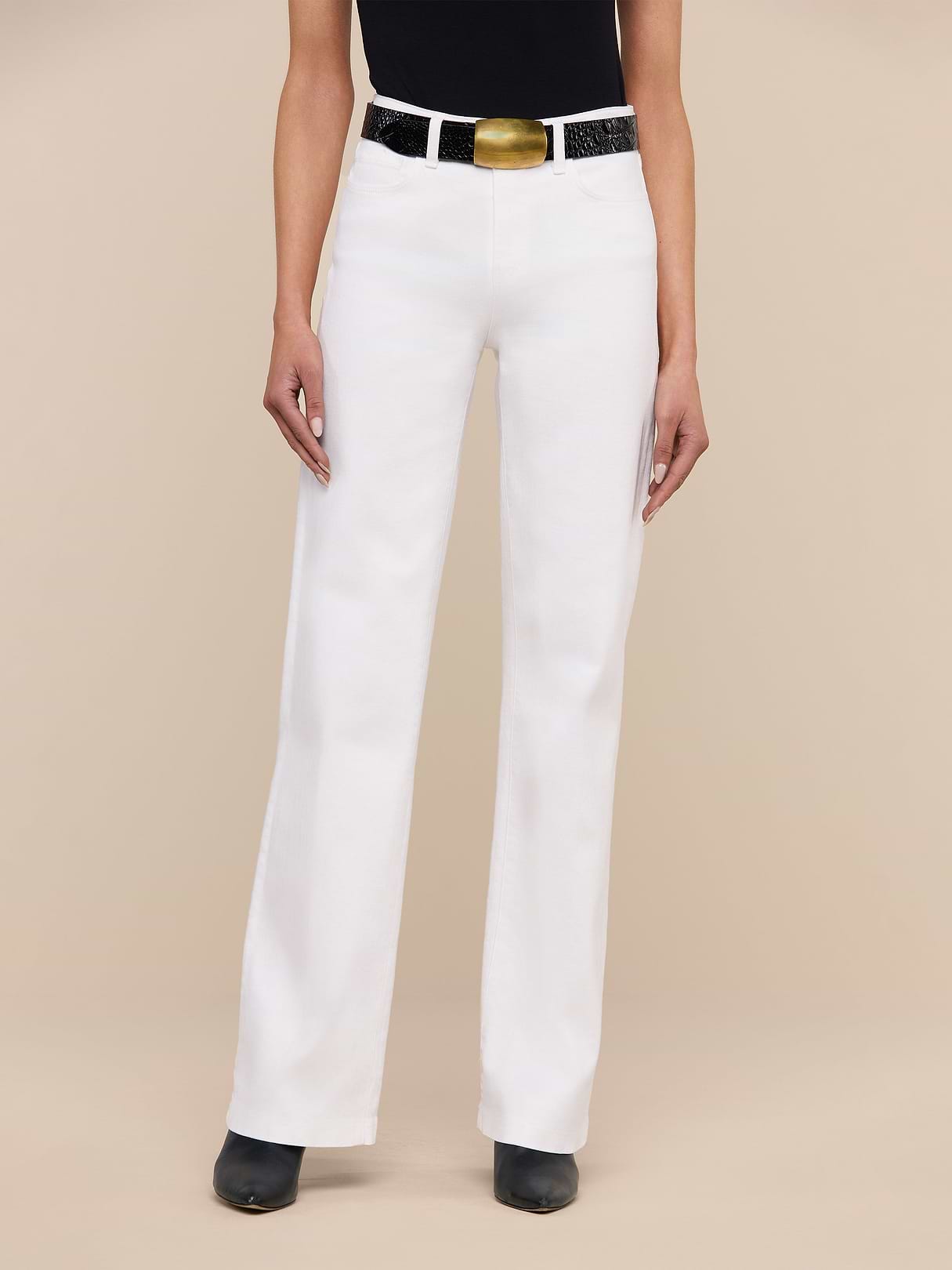 A person stands against a beige background wearing the Clayton Wide-Leg Jean in white stretch denim, styled with a black belt with a large gold buckle, a black top, and black pointed-toe shoes.