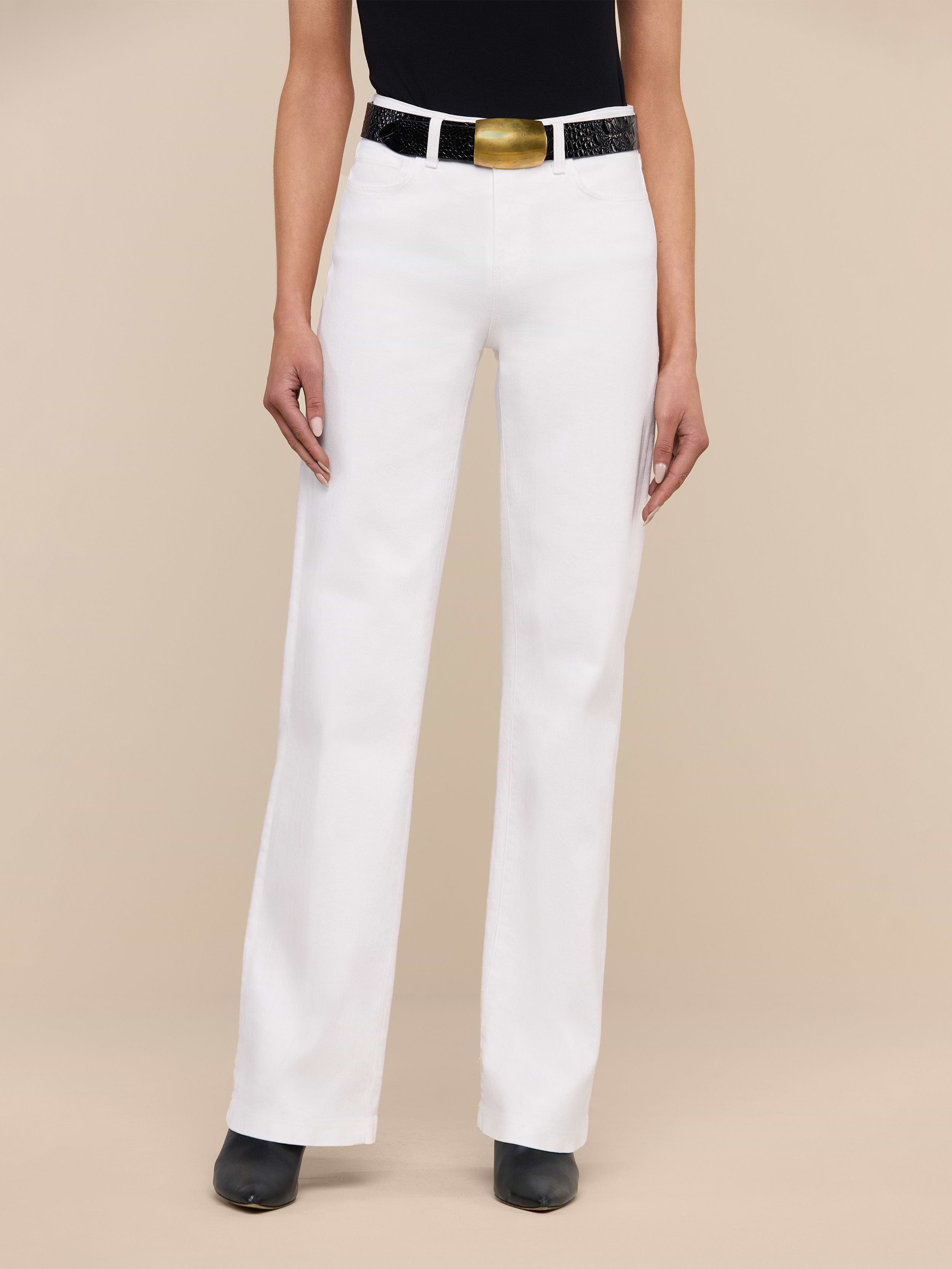 Wearing the Clayton Wide-Leg Jean in white stretch denim, paired with a black top, black boots, and a black belt featuring a large gold buckle, this person stands against a plain white background.