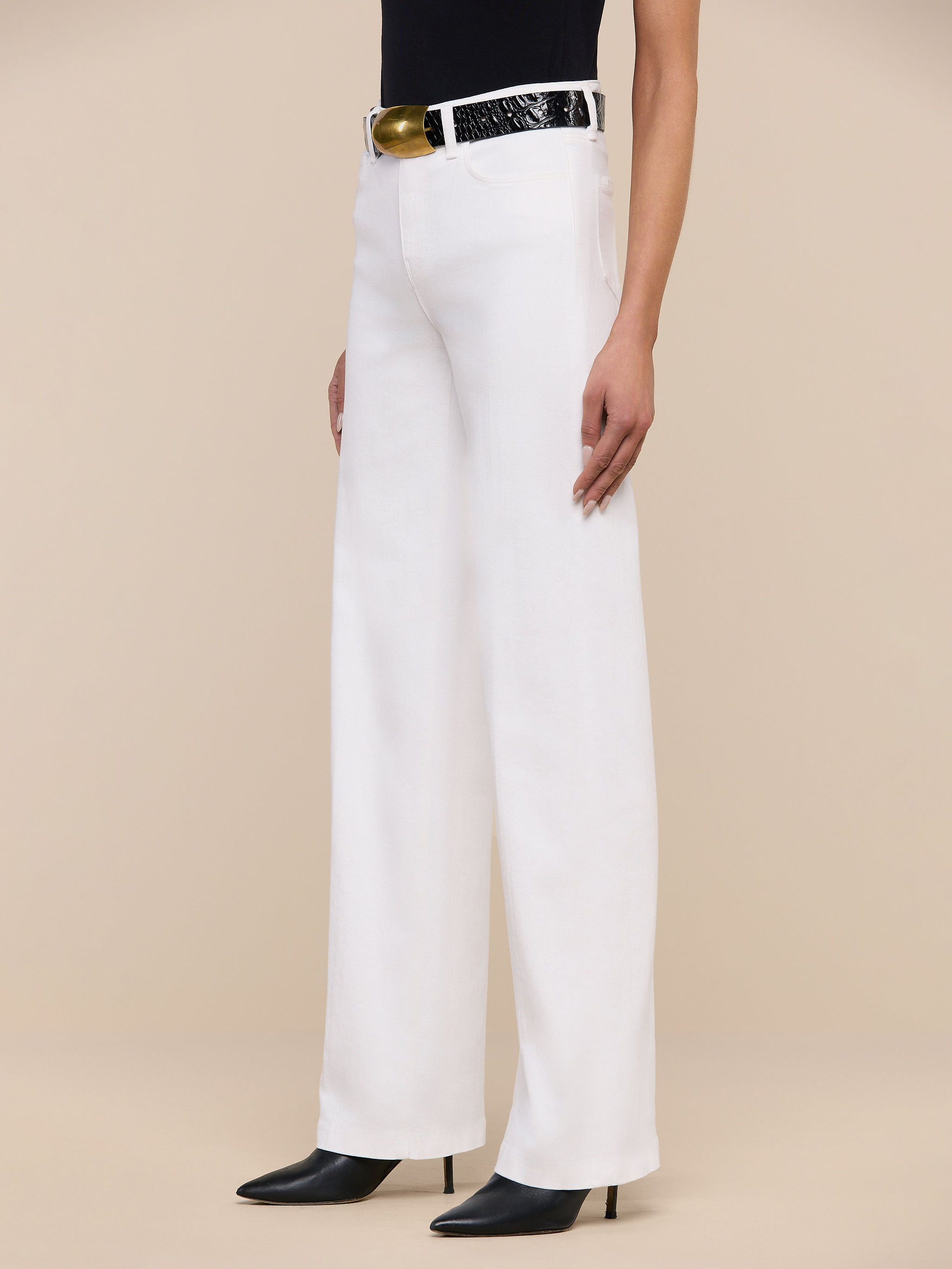A person wearing the Clayton Wide-Leg Jean in white stretch denim, paired with a black top, a black belt with a gold buckle, and black pointed-toe shoes, stands against a plain white background.