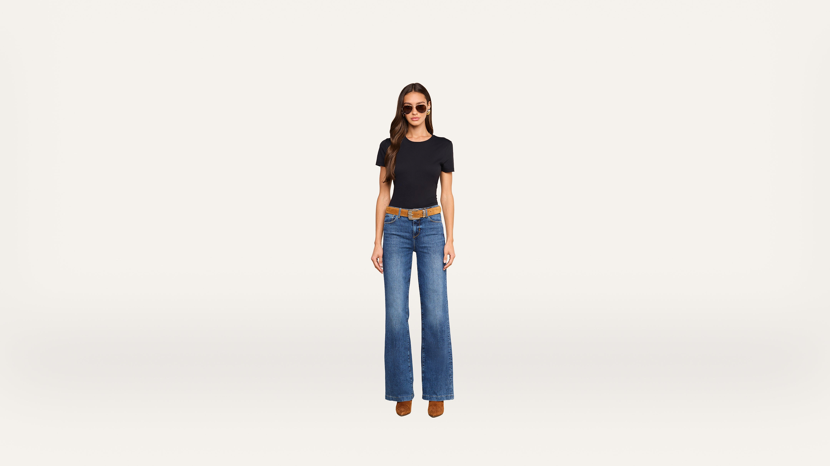 A woman wearing sunglasses, a black fitted t-shirt, wide-leg blue jeans, a brown belt, and brown shoes stands against a plain light background.