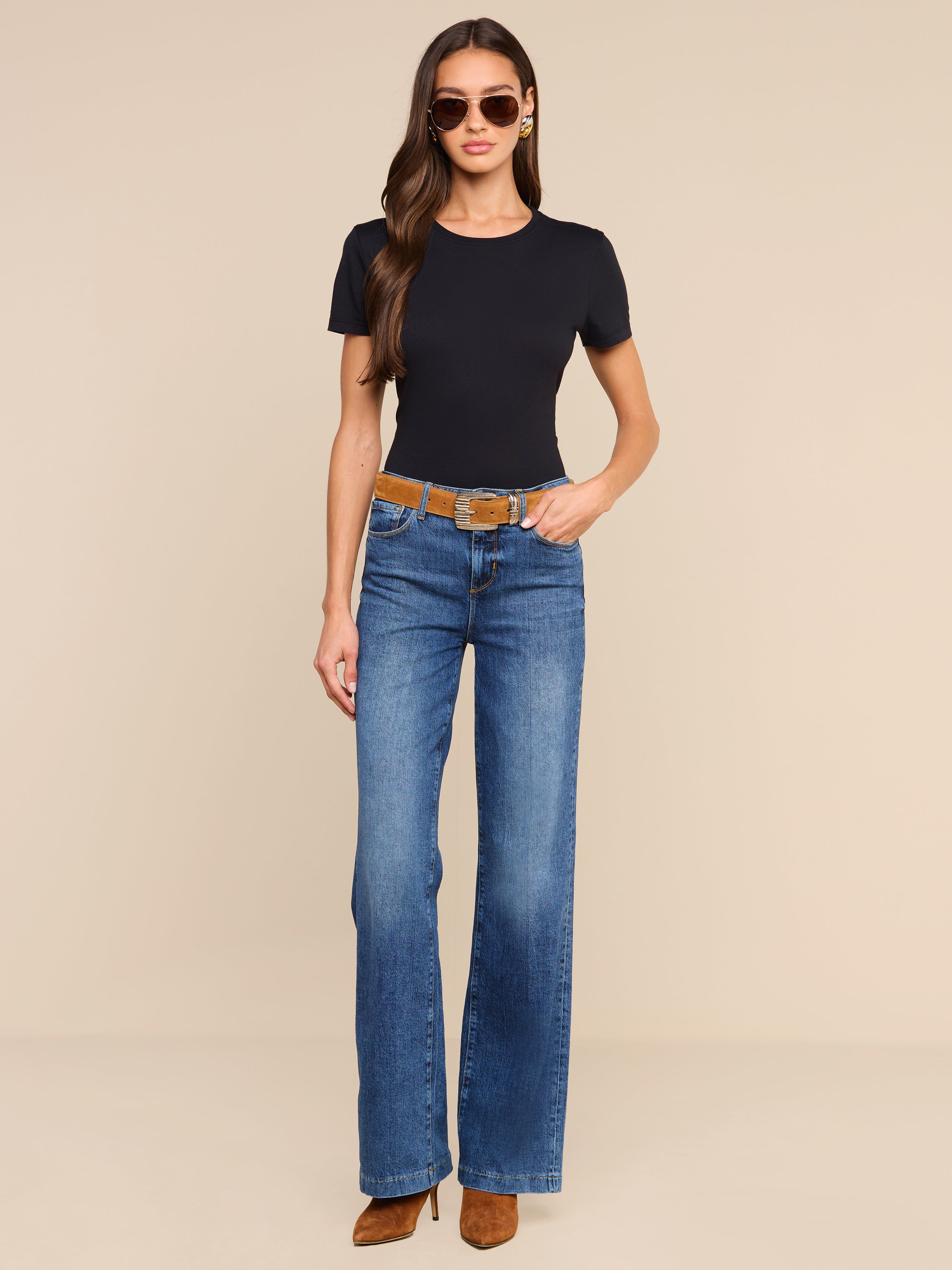 Una donna è in piedi su uno sfondo beige; indossa occhiali da sole, una maglietta nera e i jeans Clayton Wide-Leg in denim elasticizzato blu a vita alta, con una cintura marrone. Completa il look con stivali marroni con tacco e lascia che i suoi lunghi capelli ricadano liberamente, mentre tiene le mani lungo i fianchi.