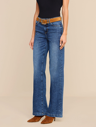 Person wearing the Clayton Wide-Leg Jean in blue high-rise stretch denim, paired with a brown belt, brown pointed-toe heeled boots, and a sleeveless black top, standing against a beige background. Only the lower half of the body is visible.