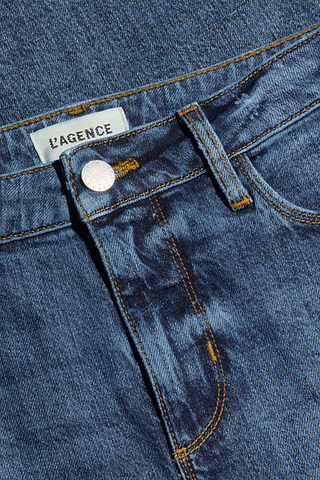 Close-up of the Clayton Wide-Leg Jean in blue denim, highlighting the waistband, metal button, belt loop, part of the pocket, and a white L’AGENCE label stitched inside. Crafted from stretch denim, the fabric showcases visible stitching and a subtly worn texture.